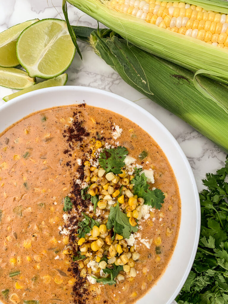 bowl of. corn chowder topped with corn, cilantro, and spices, on a marble slab next to an ear of shucked corn, lime wedges, and fresh cilantro.